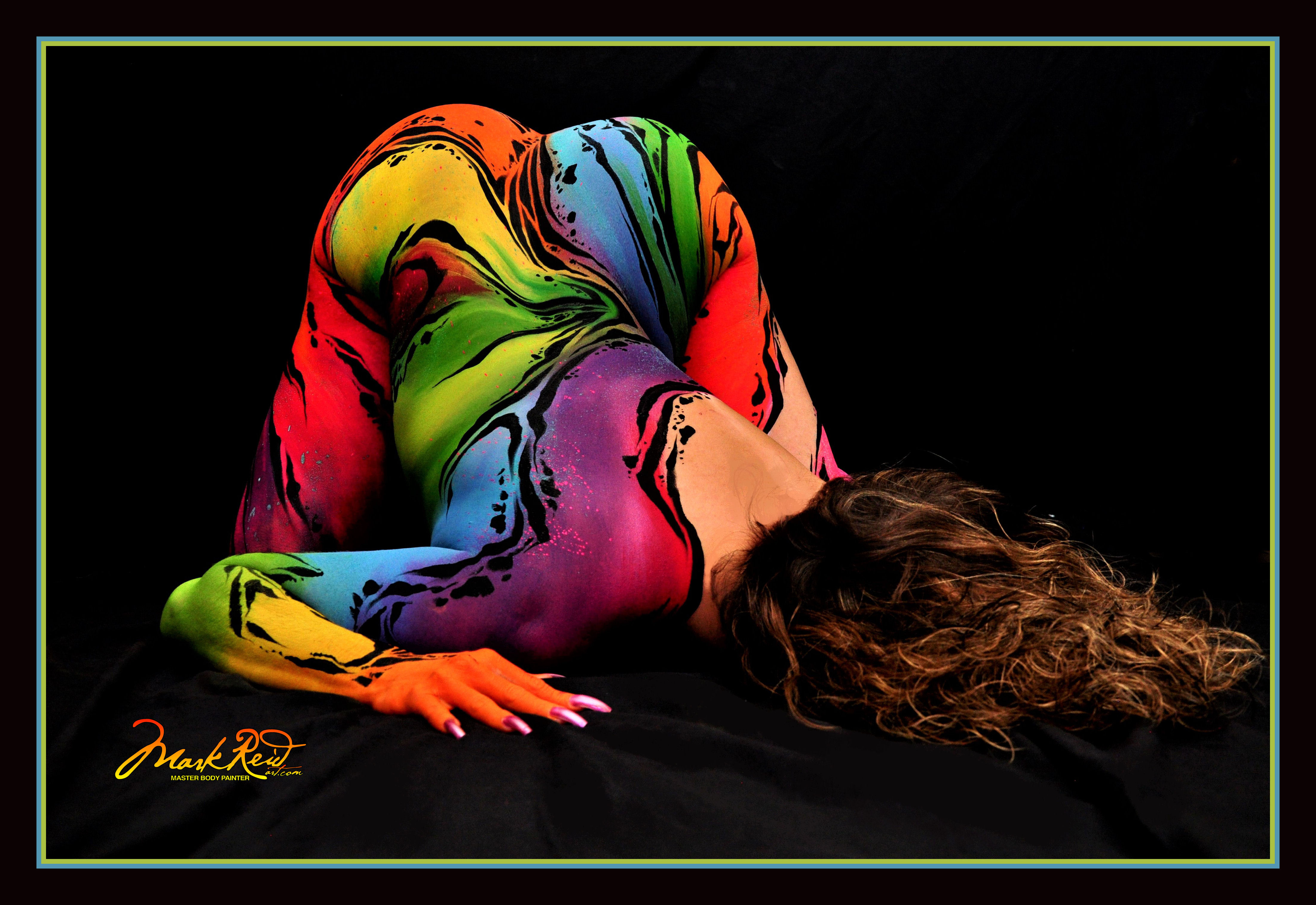 Brunette woman face down toward the camera with body paint that is brightly colored on her visible skin including her back, arms, and her buttocks.